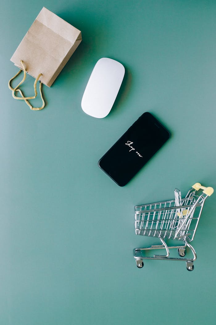 about-us Top view of a smartphone, mouse, paper bag, and mini shopping cart on a green background, symbolizing e-commerce.
